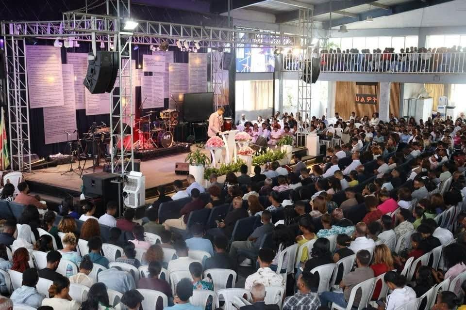 A congregation seated facing a speaker and a church stage.